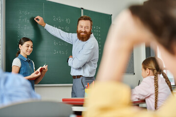 A male teacher stands before a blackboard in a vibrant classroom, instructing a group of children with enthusiasm and expertise.