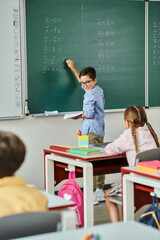 A diverse group of children sit attentively at desks in front of a blackboard, listening intently...