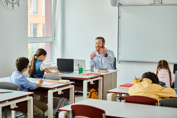 A male teacher sits before a group of students in a bright, lively classroom setting, engaging in...