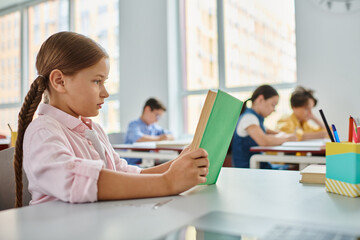 A young girl sits at a table, engrossed in a book in a bright classroom setting.