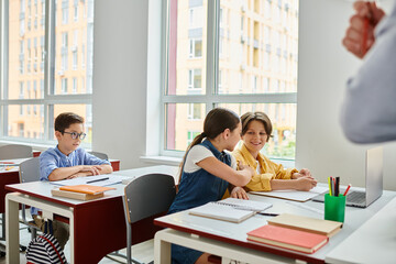 A group of children sit at desks in a bright classroom, focusing on a man teacher giving instructions.