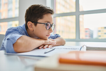 A focused young boy wearing glasses sitting attentively at a desk, engrossed in studying or working...