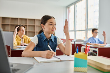 A young girl with a curious expression sits at a desk in front of a laptop computer, fully engaged in learning and exploring.