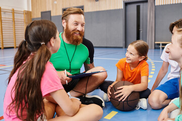 A man sits on the floor surrounded by a group of kids, teaching with enthusiasm in a colorful...