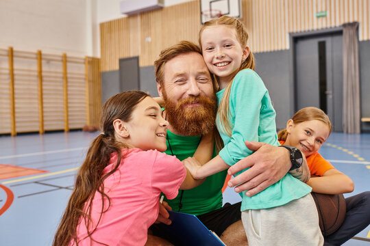 A bearded teacher with open arms, warmly hugging a diverse group of children in a vibrant classroom setting filled with laughter and joy. - Powered by Adobe