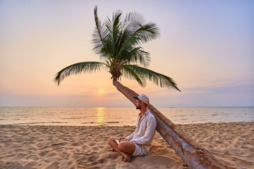 Young nomad man freelancer sitting under a palm tree and working remotely online at a laptop,...