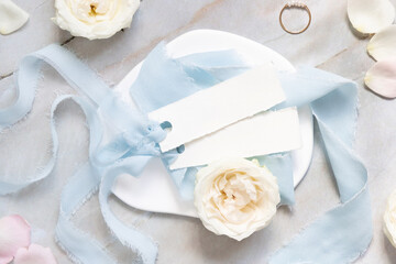 Cards near light blue fabric and cream flowers on plate top view, copy space, wedding mockup