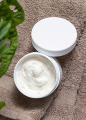  Opened cream jar with blank lid on brown towel, near green plant top view, mockup