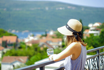 Fototapeta premium A female tourist enjoys the view of the sea coast, standing on the balcony. A girl in a hat with a glass in her hands against the backdrop of houses, sea and mountains - photo without the model’s face