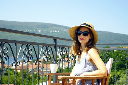 Morning coffee on the balcony of a seaside hotel (Djenovici, Montenegro). Portrait of a young smiling woman in a hat sitting on a sunny summer day on a balcony with a beautiful Mediterranean view