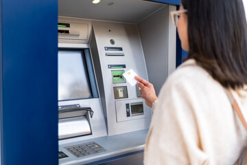 A woman uses an ATM machine, inserting her card for a transaction. The focus is on the card and machine. Finance and technology concept.