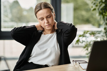 Middle-aged woman with neck pain, stressed, sitting in front of laptop.