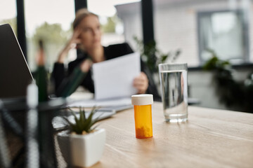 Stressed woman sits at desk with bottle of medicine.
