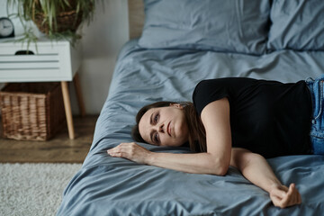 A middle-aged woman lies on her bed with arms crossed, lost in thought.