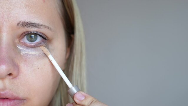 Close up of a young caucasian blonde woman applying the concealer for dark eyes. Bruises, dark circles under the eyes. Foundation cream hiding spot imperfections on female face