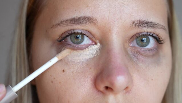 Young caucasian blonde woman applying the concealer for dark eyes. Bruises, dark circles under her eyes. Foundation cream hiding spot imperfections on female face