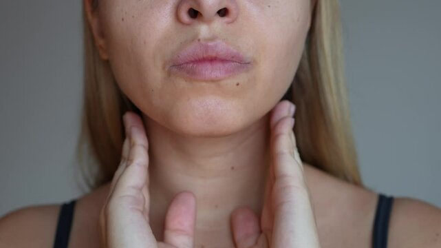 Young blonde woman with throat pain touching enlarged inflamed lymph nodes under jaw on her neck on a dark grey background. Flu, cold, covid, tonsillitis, sars virus, inflamed tonsils. Sore throat