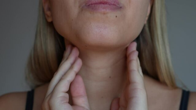 Young blonde woman with throat pain touching enlarged inflamed lymph nodes under jaw on her neck on a dark grey background. Flu, cold, tonsillitis, inflamed tonsils. Sore throat. It hurts to swallow