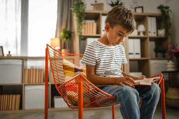 young small caucasian boy sit on the orange chair and read a book