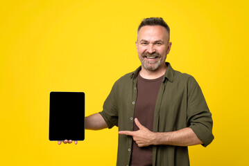 Smiling middle-age mature man pointing to his tablet pc with black screen mock-up on yellow background with thumb-up gesture. Handsome man dressed in green shirt holding big tablet pc in hand