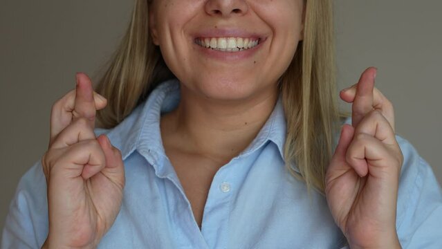 Close up of a young happy caucasian smiling blonde woman in a blue shirt crossing her fingers for good luck waiting for the results of the lottery or exams on a grey background