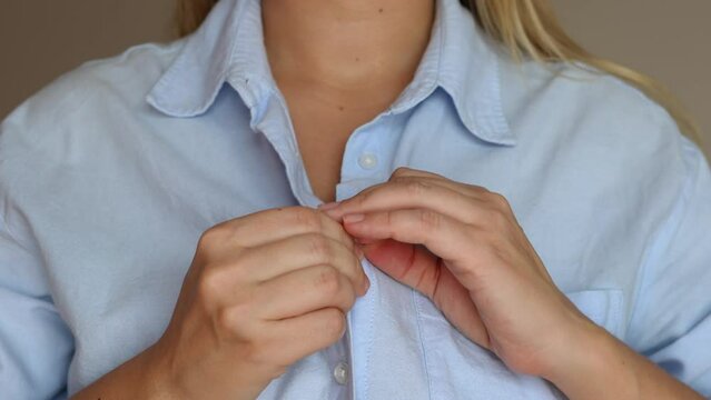 Cropped shot of a young blonde woman in a blue shirt buttoning the collar on a gray background