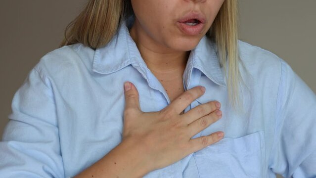 Cropped shot of a young blonde woman keeping her hand on her chest in pain taking deep breaths of air on a grey background. Chest pain, lack of breath, panic attack