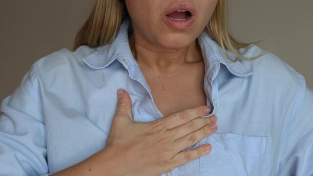 Cropped shot of young blonde woman keeping her hand on her chest in pain taking deep breaths of air unbuttoning the top button of her shirt on grey background. Chest pain, lack of breath, panic attack