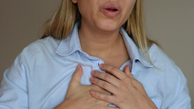 Cropped shot of a young blonde woman keeping her hand on her chest in pain taking deep breaths of air on a grey background. Chest pain, lack of breath, panic attack. Slow motion