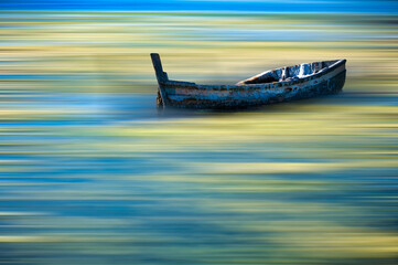 Fototapeta premium An old, weathered boat floats serenely on water with vibrant, blurred streaks of blue and yellow hues.