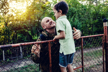 An emotional military father, dressed in camouflage, holds his young son in arms in greeting after returning home. Little boy meeting his military father at home.