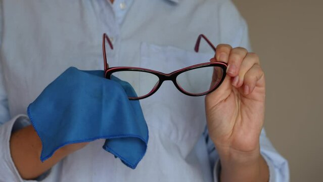 Cropped shot of a young woman wiping her glasses with a blue cloth on beige background. Fashionable glasses for working at a computer with a blue filter lenses. Anti blue light and rays