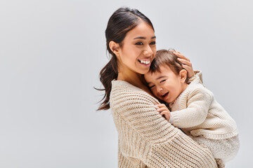 A young Asian mother tenderly embracing her child in a studio setting against a grey background.