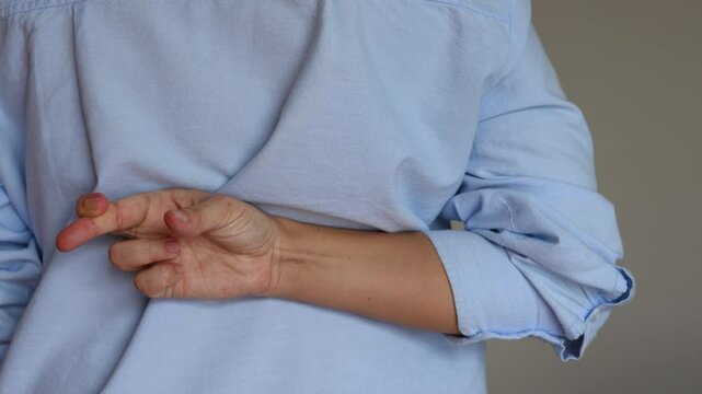 Cropped shot of a young woman in a blue shirt crossing her fingers behind her back on a grey background. Deception, lies, cheat
