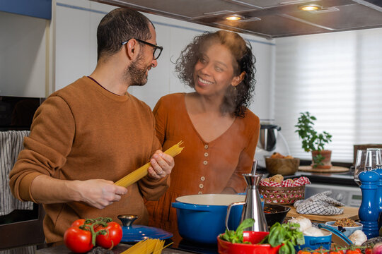 A Hispanic couple cooking spaghetti together in the cozy kitchen of their home while looking at each other in love and smiling - Powered by Adobe