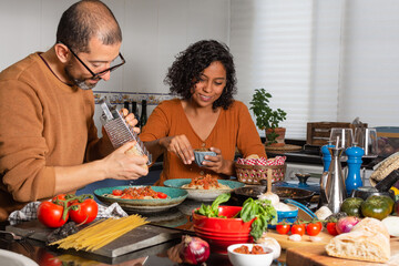 Young Hispanic couple in the kitchen preparing food. Man grates cheese while woman puts seasoning on delicious pasta