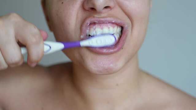 Cropped shot of a young caucasian woman brushing her teeth with a toothbrush in a circular motion on a grey background. Morning and evening routine. Close up