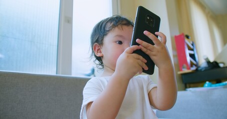 Adorable toddler child Playing engrossed in a smartphone while sitting on a sofa in a bright,...