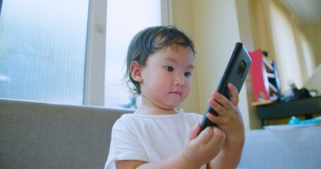 Adorable toddler child Playing engrossed in a smartphone while sitting on a sofa in a bright,...