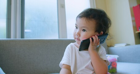 Cute Toddler child holding a smartphone to their ear Talking on Smartphone While Sitting on Couch in Bright Living Room expressing surprise.