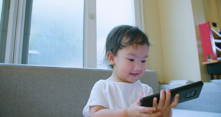 Adorable toddler child Playing engrossed in a smartphone while sitting on a sofa in a bright,...