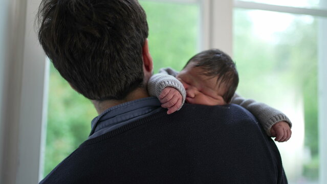 Newborn baby resting on the father’s shoulder, dressed in a cozy knitted sweater. The father turns body towards window, highlighting the comforting and secure embrace between them