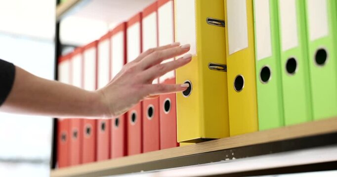 Secretary puts yellow folder with materials back on shelf of rack in office. Organized multi-colored ring binders with archive files slow motion