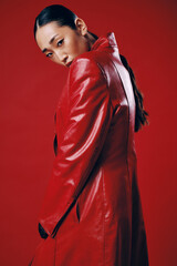 Fashionable asian brunette woman in a red leather jacket posing with hands on hips against red...