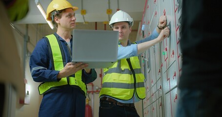 Two electricians electrical engineers in protective uniform checking voltage control panel screen system at electrical cabinet for generate electricity of factory in manufacture industrial