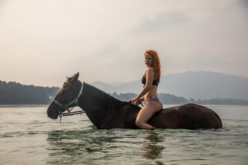 A beautiful girl stands on a horse in the water. Brown horse. Girl with red hair. Bathing horses.	