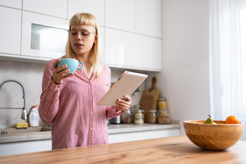 Young businesswoman real estate agent morning routine, drinking coffee in kitchen using digital tablet to read emails. Working at home female professional designer, business owner, confident lifestyle