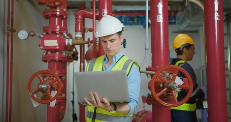 Engineer in safety gear inspecting red industrial pipes while holding using laptop in a factory environment, ensuring proper maintenance and functionality.