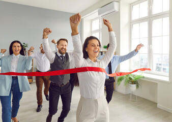 Happy woman employee running to the red ribbon with raised arms celebrating crossing finishing line...