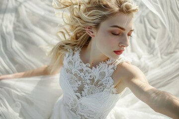 Beautiful young bride with long blonde hair blowing in the wind is wearing a white lace wedding dress and posing on white fabric
